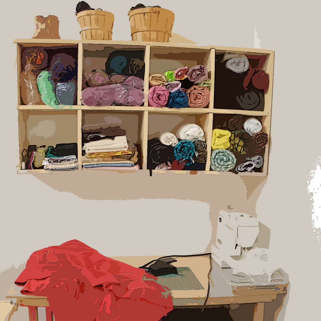 system, filled with rolls of fabric. Atop are three baskets, each one larger than the one to the left. Below the cubby is the top of a desk, on which sits a white sewing machine on the right. The eye follows a wire from the back of the sewing machine to the pedal, also sitting on the desk, on top of a green cutting mat. To the immediate left lies a large red swath of fabric, it dangles over the front and left edges of the table.